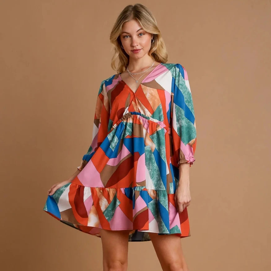 A blonde woman modeling a long-sleeve geometric print mini dress with a tiered ruffle hem against a neutral background.