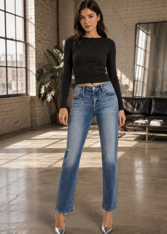 A woman modeling a pair of medium-wash blue straight-leg jeans with a raw hem, paired with a black long-sleeve crop top and silver pointed heels in a bright loft-style room with brick walls and large windows.