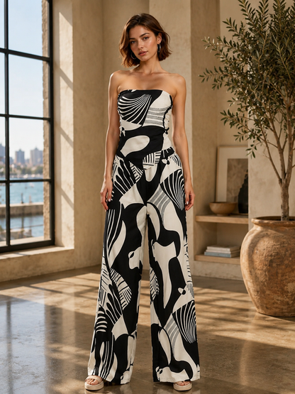 A woman models a strapless wide-leg jumpsuit with a bold black and off-white abstract pattern in a sunlit indoor room with large windows and a potted plant.