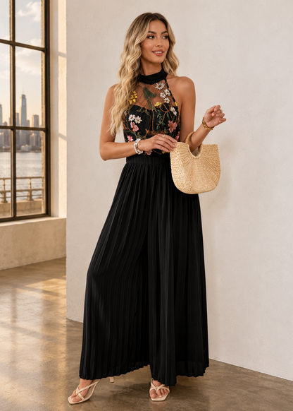 A woman models a sleeveless black pleated maxi dress with a floral embroidered bodice and sheer mesh neckline. She is holding a woven straw tote bag and wearing beige strappy heels, standing in a bright room with a window showing a city skyline.