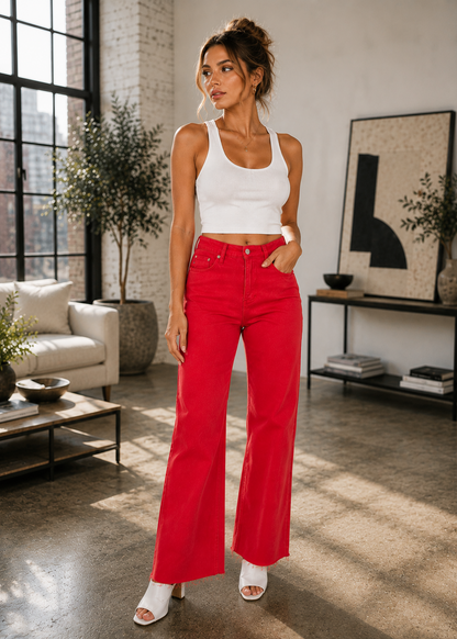A woman stands in a bright, modern loft-style room wearing a white ribbed tank top and bright red wide-leg jeans, paired with white heeled sandals.