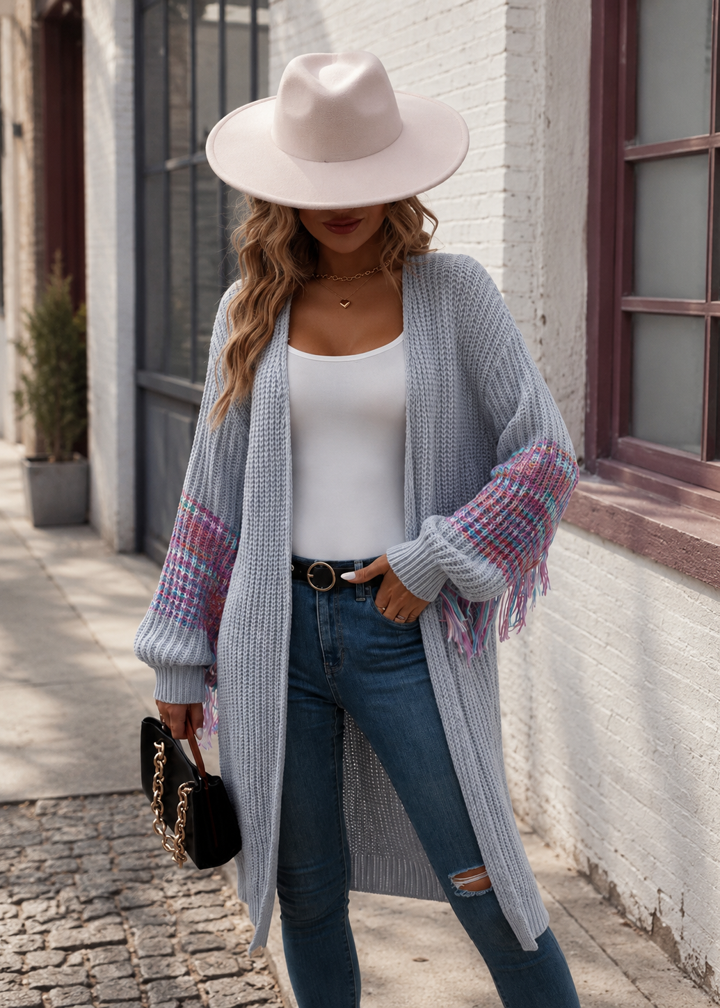 A woman stands on a cobblestone street wearing a light gray waffle-knit open-front cardigan with pink and blue plaid sleeves and fringe, a white tank top, blue jeans, and a wide-brimmed light pink hat.