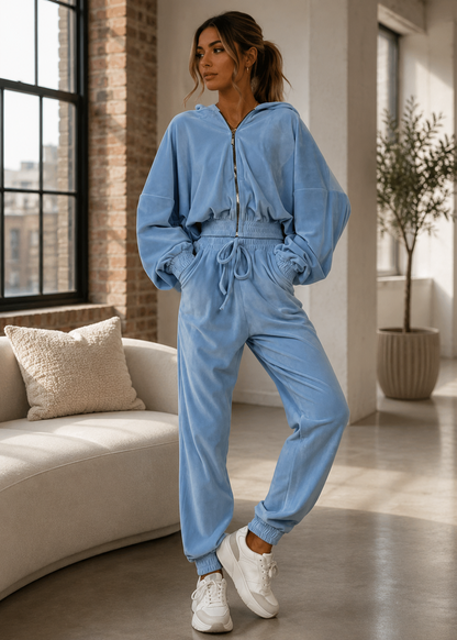A woman modeling a light blue zip-up hooded sweatshirt and matching jogger pants set, styled with white sneakers in a modern living room setting.