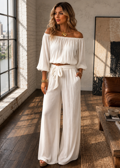 A woman models a two-piece white off-the-shoulder crop top and matching wide-leg pants set in a sunlit room with a large window, wooden floor, and brown leather chair.
