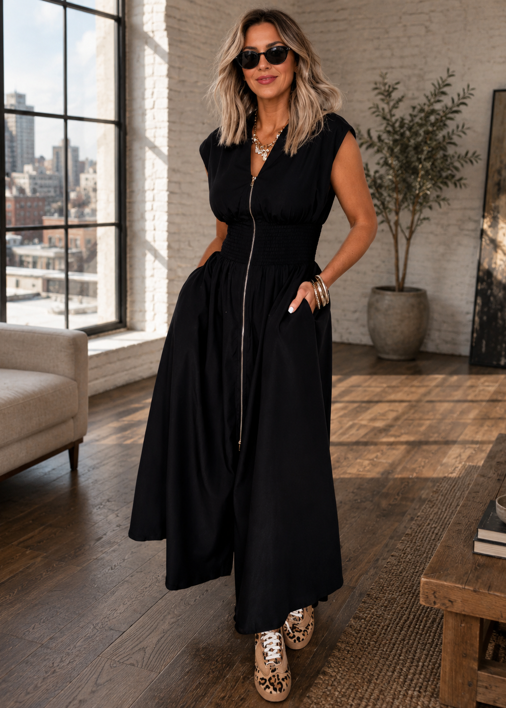 A woman wearing a sleeveless black midi dress with a front zipper and side pockets, styled with gold sneakers and sunglasses in a modern loft-style room with a brick wall and large window.