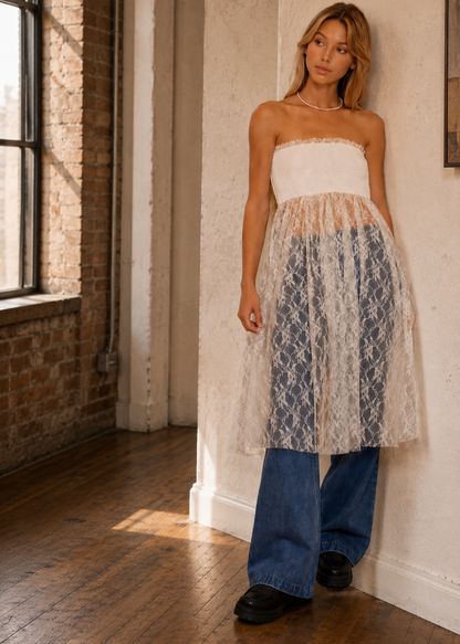 A woman stands in a sunlit loft room wearing a strapless white tube top and a sheer, layered midi skirt with a blue and white geometric lace pattern, styled with wide-leg blue jeans and black shoes.