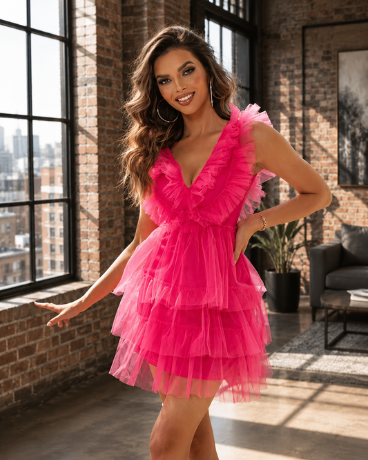A woman models a bright pink ruffled mini dress in a loft-style room with brick walls and large windows.