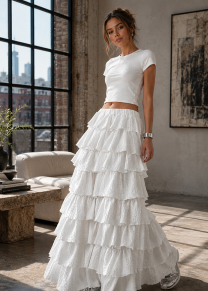 A woman models a white two-piece outfit consisting of a short-sleeve crewneck crop top and a long, tiered ruffle maxi skirt, standing in a modern loft-style room with large windows.