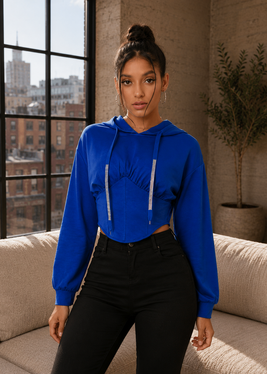 A woman wearing a bright royal blue cropped hoodie with silver drawstrings and black pants, standing in a modern loft setting with a window and beige couch.