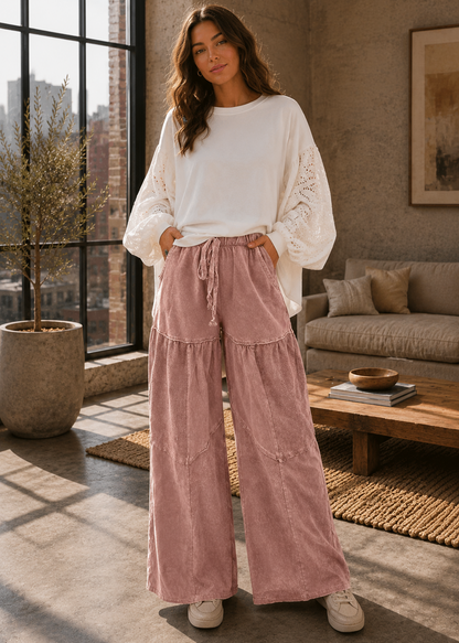 A woman stands in a sunlit living room wearing a white long-sleeve top with lace-up sleeves and dusty pink wide-leg pleated pants. She is posed in front of a large window, with a sofa, coffee table, and plant visible in the background.