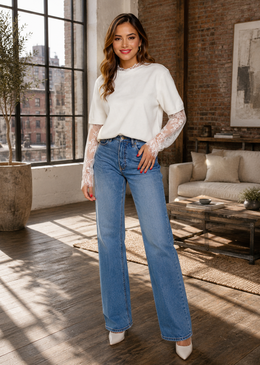 A woman stands in a bright loft-style room wearing a white long-sleeve top with sheer lace sleeves and light blue wide-leg jeans, paired with white pointed heels.
