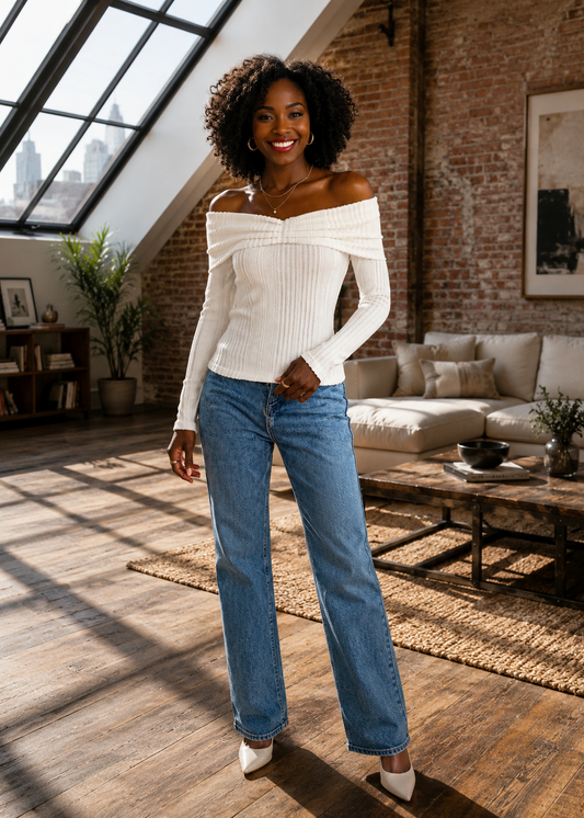 A woman stands in a sunlit loft apartment wearing a ribbed off-the-shoulder cream top and blue wide-leg jeans, with a brick wall, sofa, and large window behind her.