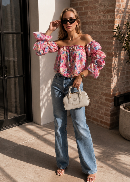 A woman stands outdoors wearing a colorful off-the-shoulder puff-sleeve floral top, light-wash wide-leg jeans, sunglasses, and carrying a structured light beige handbag.