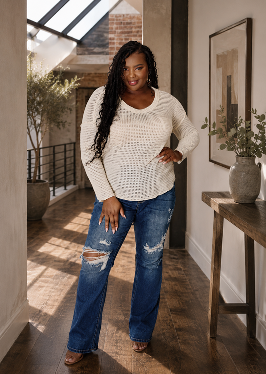 A woman modeling a cream-colored textured knit sweater and distressed blue jeans in a modern loft hallway with wooden floors and a skylight.