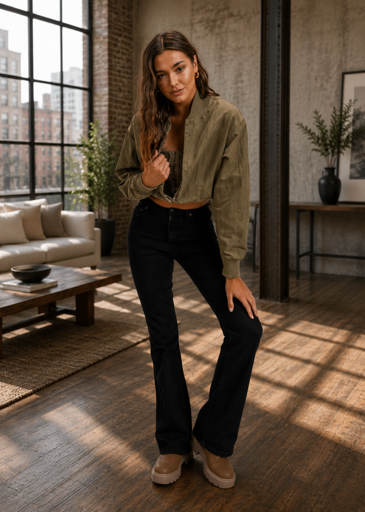 A woman stands in a sunlit loft apartment wearing an olive green cropped button-up shirt, black high-waisted flare jeans, and beige slip-on shoes.