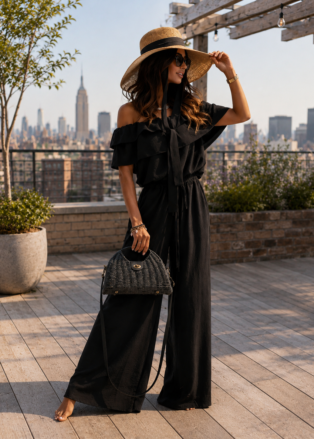 A woman stands on a rooftop terrace wearing a black off-the-shoulder wide-leg jumpsuit, a wide-brimmed straw hat, and carrying a black quilted handbag, with a city skyline in the background.