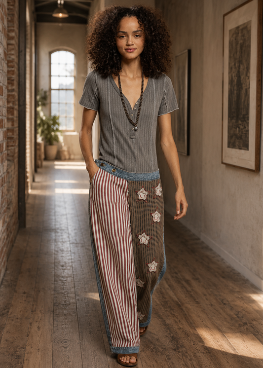 A woman walking down a sunlit hallway wearing a gray striped short-sleeve top and wide-leg pants with red-and-white vertical stripes and embroidered details.