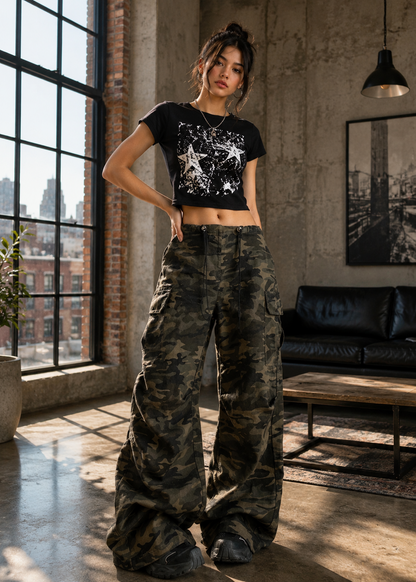 A woman stands in a loft-style room with large windows, concrete walls, and a black leather sofa, wearing a black cropped graphic T-shirt and very wide-leg camouflage cargo pants.