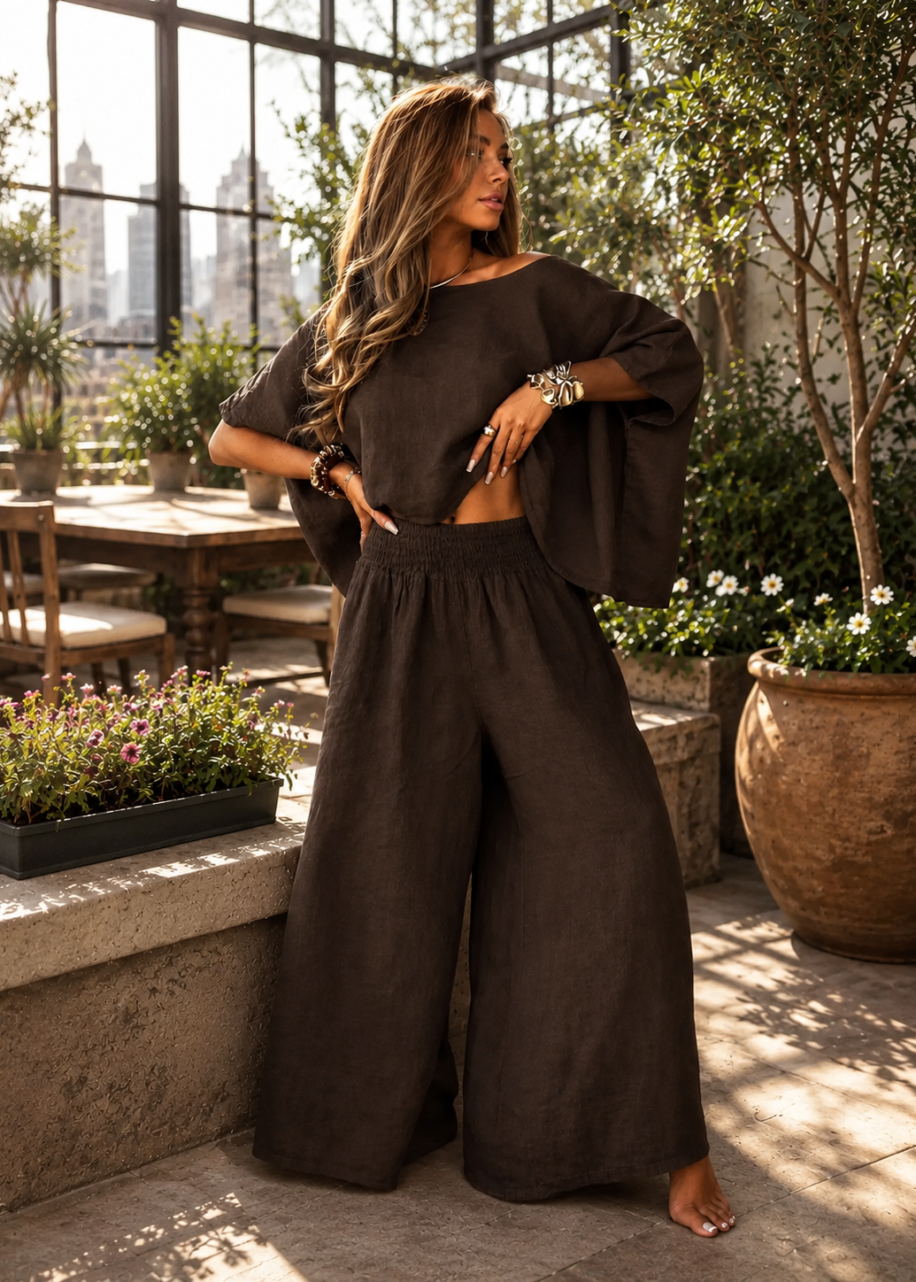 A woman models a dark brown two-piece linen set consisting of a short-sleeve crop top with wide flared sleeves and matching high-waisted, wide-leg pants, set in a sunlit patio environment with potted plants.
