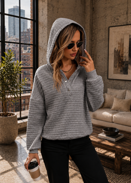 A woman wearing a light gray waffle-knit hooded pullover sweater with a partial button placket, styled with black pants in a modern loft setting with a window, sofa, and coffee table.