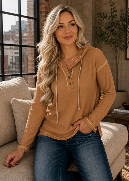 A woman with long blonde hair sits on a beige couch wearing a tan long-sleeve hooded henley top with drawstrings and blue jeans in a bright living room setting.