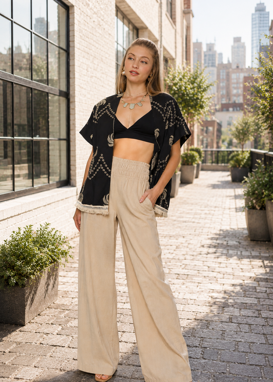 A woman stands on a cobblestone walkway in an urban setting, wearing a black cropped open-front cardigan with gold geometric embroidery over a black bandeau top and high-waisted beige wide-leg pants.