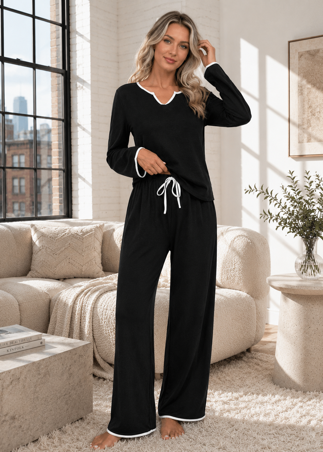 A woman modeling a black two-piece pajama set with white trim, consisting of a long-sleeve V-neck top and matching wide-leg pants, standing in a bright living room.