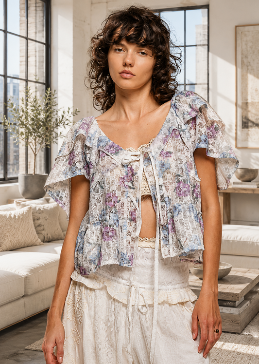 A woman modeling a bohemian-style short-sleeve crop top with a purple and blue floral print on a white background, paired with cream lace pants in a bright, modern loft living room.