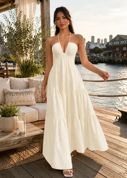 A woman stands on a wooden dock by the water wearing a sleeveless, tiered maxi dress in a warm off-white or cream color. The background features a patio with cushions, potted plants, and a waterfront view with a sign reading 'Red Hook Dock'.