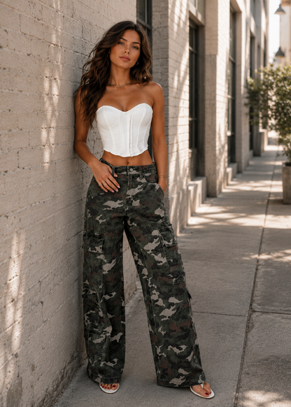 A woman stands on a city sidewalk against a light brick wall, wearing a white strapless corset-style top and loose camouflage cargo pants with sandals.
