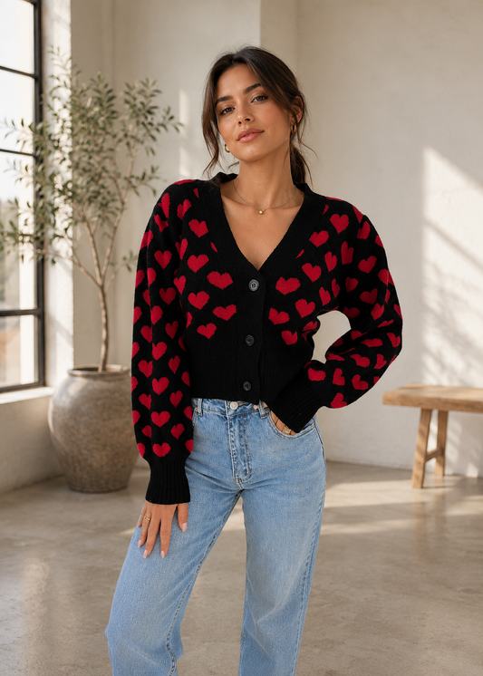 A woman modeling a black cardigan sweater with a red heart pattern, paired with light blue jeans in a bright, modern room with a window and potted plant.