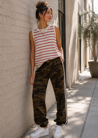 A woman stands on a sunlit sidewalk against a light brick wall, wearing a sleeveless red-and-white striped top, olive-brown camouflage jogger pants with white side stripes, and white sneakers.