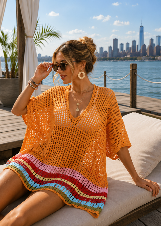 A woman sits on a white lounge chair by a waterfront wearing a bright orange crochet cover-up dress with multicolored striped trim. The background shows a city skyline across the water under a blue sky.