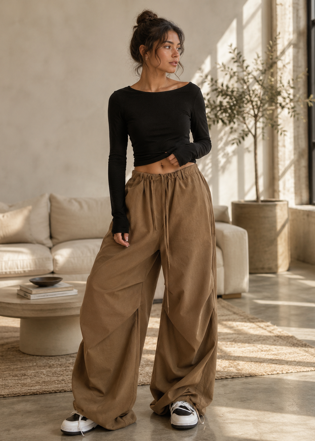 A woman stands in a sunlit living room wearing a fitted black long-sleeve crop top and very loose, wide-leg brown pants with white sneakers.