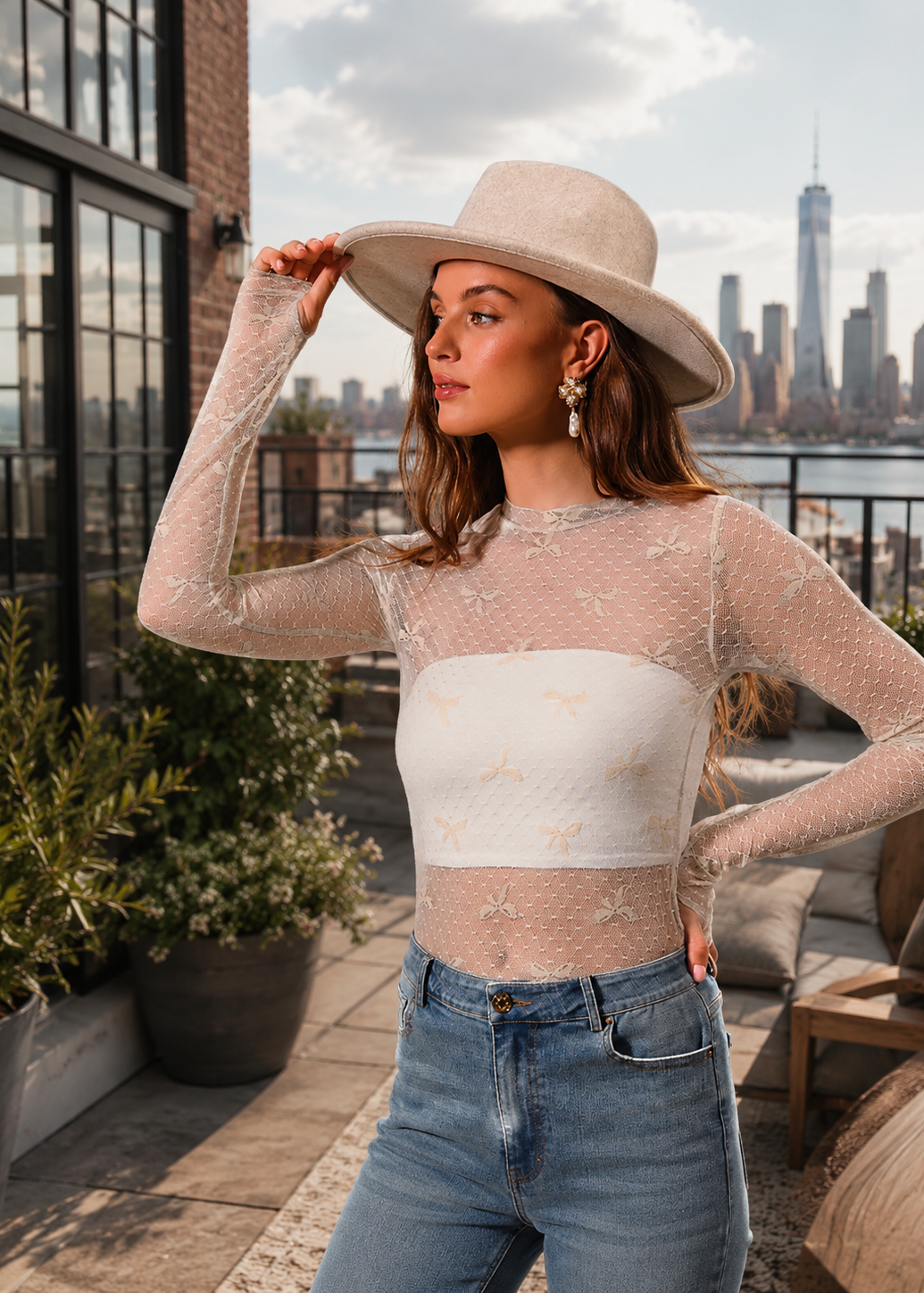 A woman wearing a beige wide-brim fedora hat and a sheer beige long-sleeve crop top with a white horizontal band, paired with blue jeans, posing on a rooftop terrace with a city skyline background.