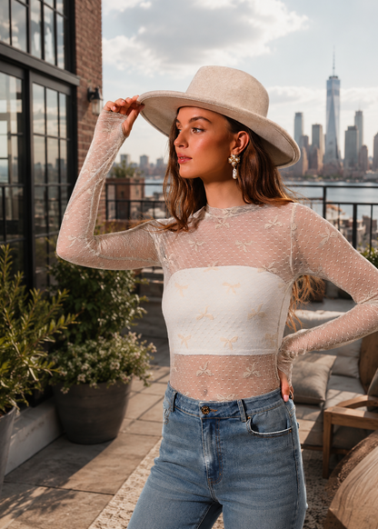 A woman wearing a beige wide-brim fedora hat and a sheer beige long-sleeve crop top with a white horizontal band, paired with blue jeans, posing on a rooftop terrace with a city skyline background.