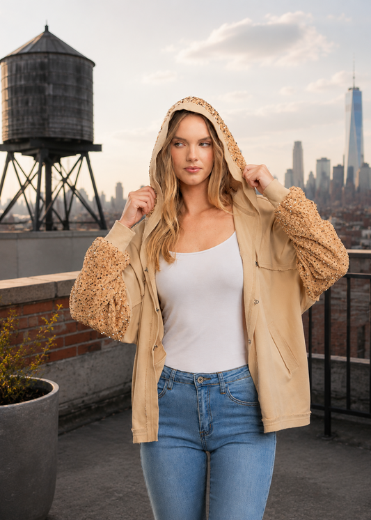 A woman stands on a rooftop wearing a beige hooded jacket with textured, fuzzy sleeves and a white tank top, with a city skyline and a water tower in the background.