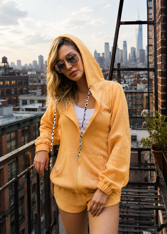 A woman wearing a fuzzy mustard yellow zip-up hoodie and matching shorts stands on a city rooftop balcony with a skyline background.