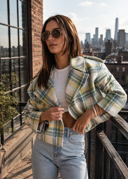 A woman wearing a cropped plaid blazer in yellow, green, and white over a white crop top and light blue jeans, standing on a balcony with a city skyline in the background.
