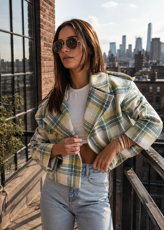 A woman wearing a cropped plaid blazer in yellow, green, and white over a white crop top and light blue jeans, standing on a balcony with a city skyline in the background.