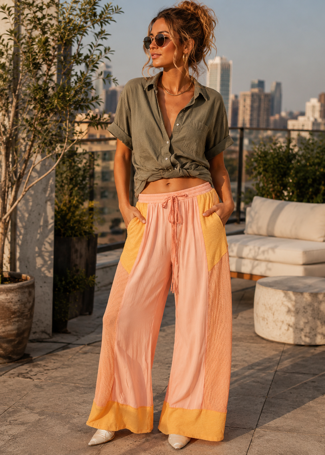 A woman stands on a rooftop terrace wearing wide-leg colorblock pants in pink and yellow, paired with an olive green tied button-up shirt. The background shows a city skyline at sunset with potted plants and outdoor seating.