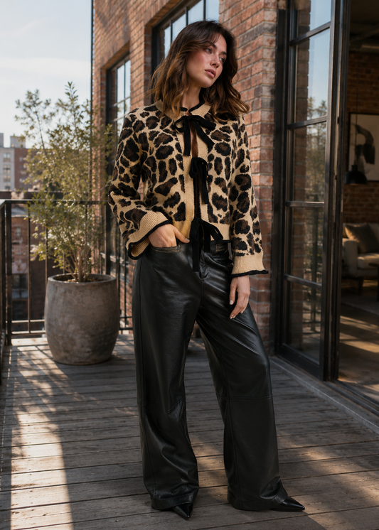 A woman stands on a wooden balcony wearing a beige and black leopard-print cropped jacket with a black bow tie-neck detail, paired with black wide-leg leather pants.