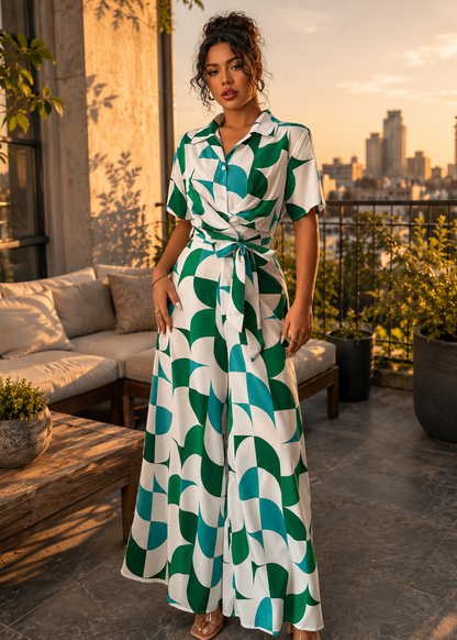 A woman stands on a rooftop terrace at sunset wearing a white short-sleeve collared dress with a bold green geometric pattern and a matching tie waist. A sofa, plants, and city skyline are visible in the background.