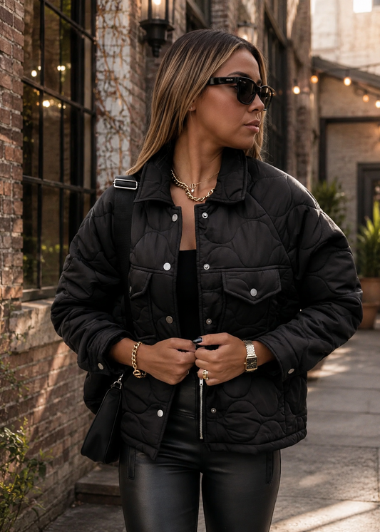 A woman wearing a black quilted puffer jacket, black sunglasses, and black leather pants, standing in an outdoor alleyway next to a brick wall.