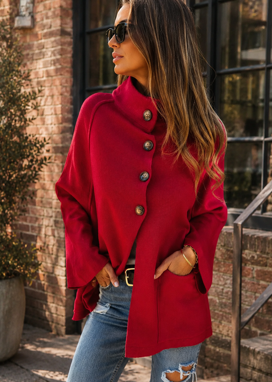 A woman wearing a bright red asymmetrical coat with large decorative buttons, styled with blue jeans and sunglasses, standing outdoors in front of a brick building.