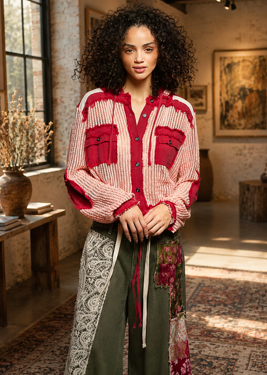 A woman stands in a rustic indoor room wearing a long-sleeve button-front shirt with red and cream vertical knit panels, red chest pockets, and a matching red tie detail, paired with olive green wide-leg pants featuring lace and floral patchwork panels.