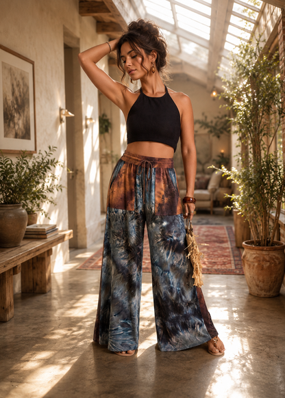 A woman stands in a sunlit indoor corridor wearing a black halter crop top and wide-leg tie-dye pants with blue, brown, and orange tones. She is holding a small tan handbag and wearing flat sandals.