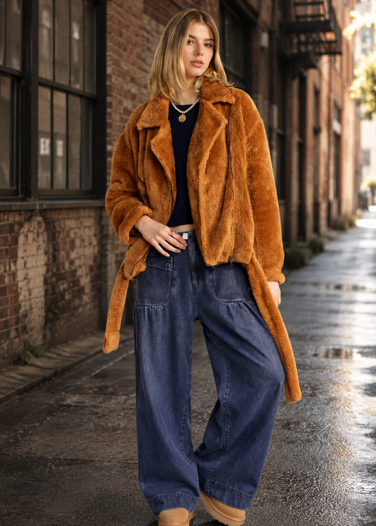 A woman stands on a wet city sidewalk wearing a camel-brown plush faux-fur coat over a dark top and wide-leg blue jeans.