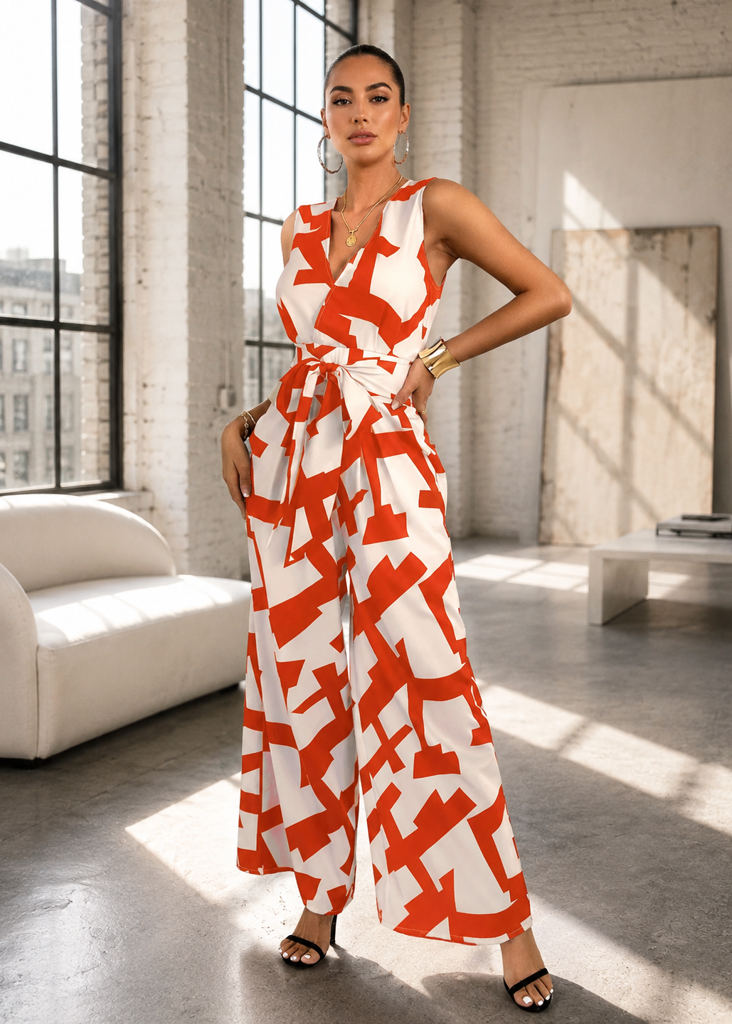 A woman models a sleeveless white jumpsuit with a bold orange geometric pattern and a matching waist tie, standing in a bright, modern loft with large windows and concrete floors.