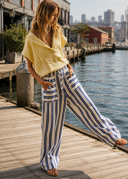 A woman stands on a wooden dock by the water wearing a pale yellow knit top and high-waisted, wide-leg striped pants with vertical navy, white, and light blue stripes. The background shows a waterfront city scene with buildings and boats.